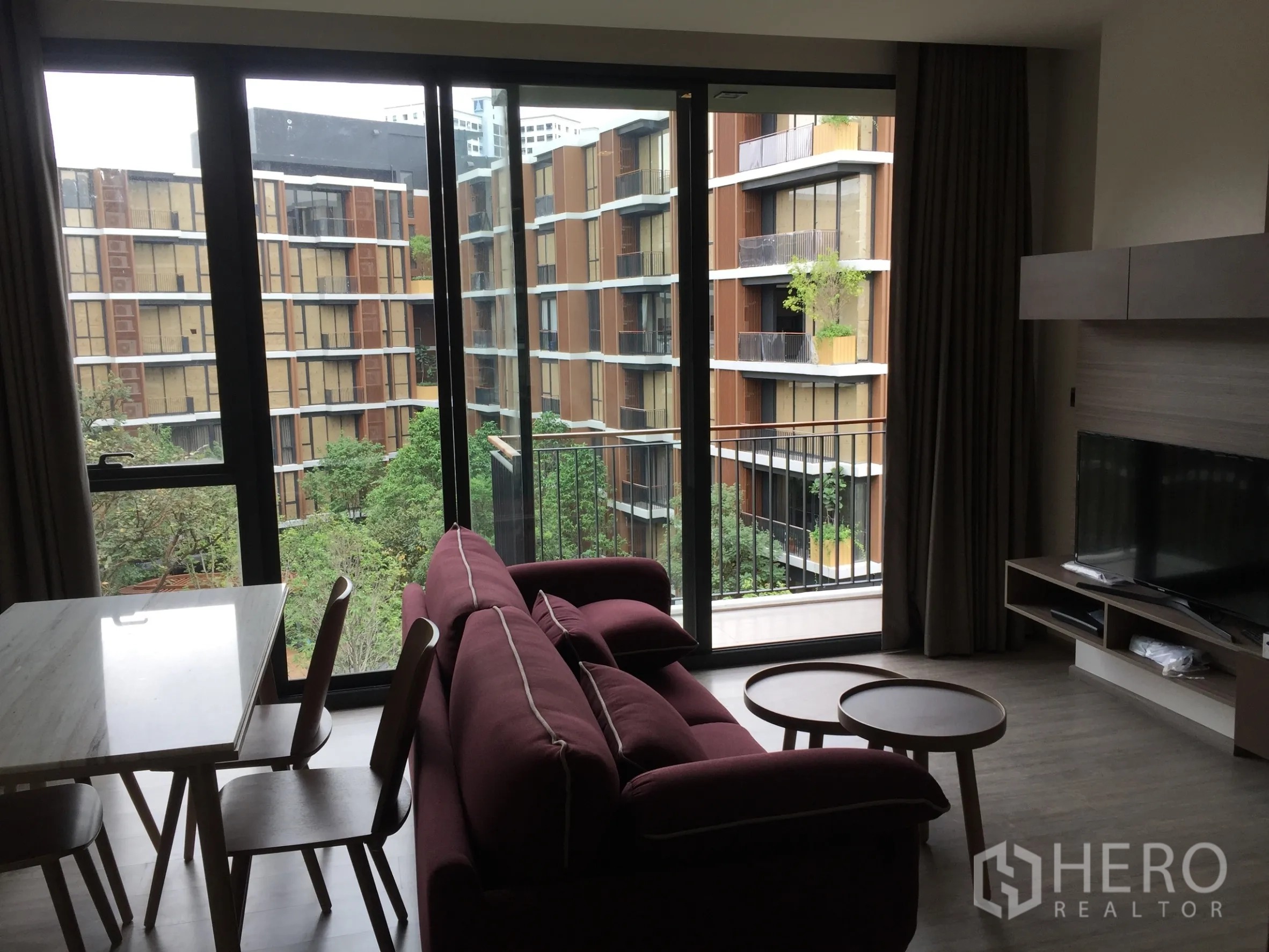 คอนโด ให้เช่าในWatthana, Bangkok - ห้องนั่งเล่นโซฟาสีแดงไวน์และทีวี เปิดสู่ระเบียงผ่านหน้าต่างสูงจรดเพดาน มองเห็นสวนส่วนกลางร่มรื่น.