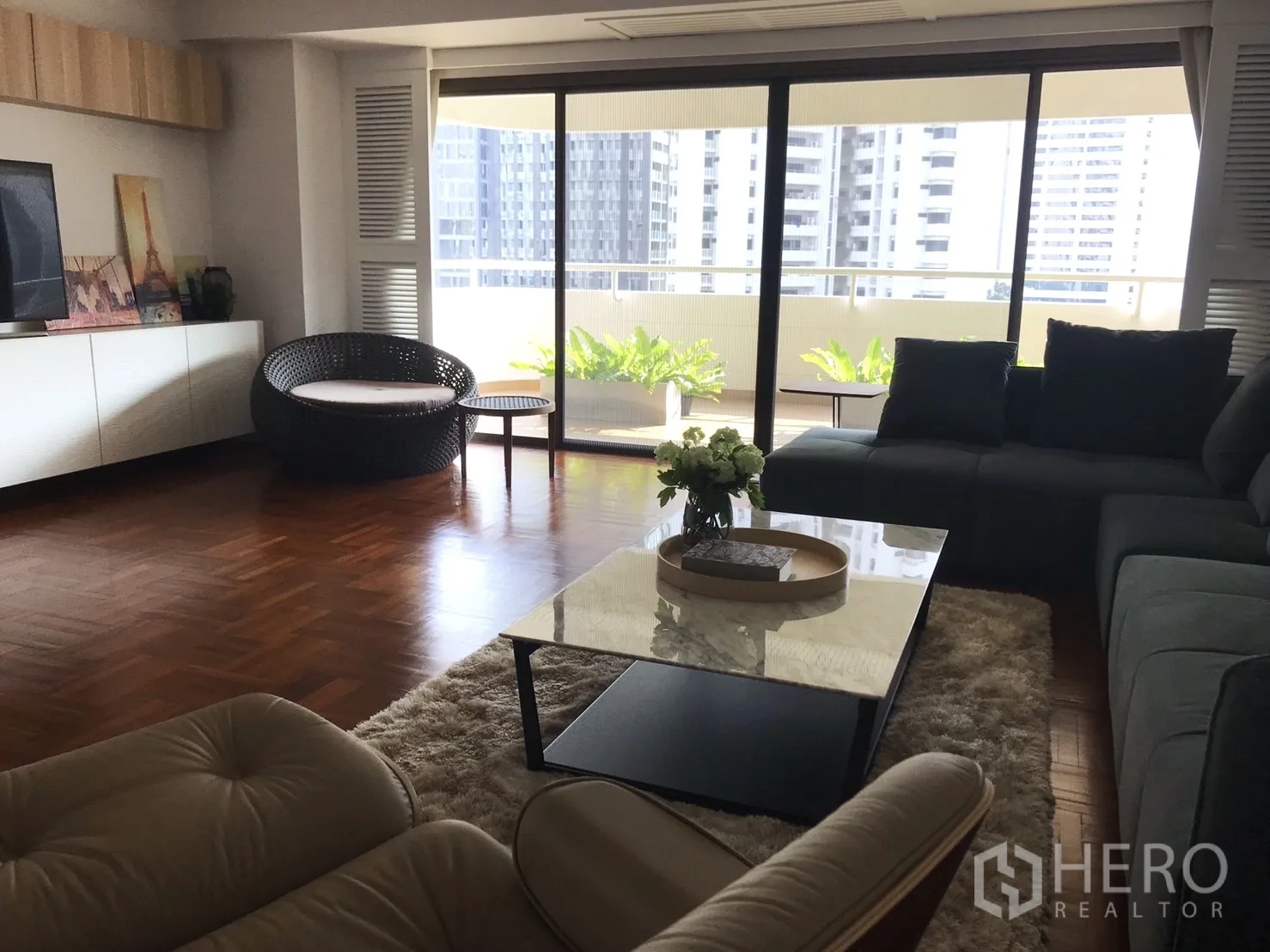 Condominium for rent in Bangkok - Bright living room with parquet floors, sectional sofa, and sliding doors opening to a wide balcony with city view and planters.