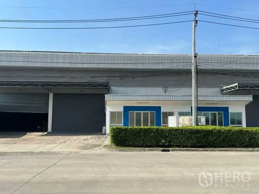 工厂 出租在Bang Sao Thong, Samut Prakan - 工厂正面，带大卷帘门与蓝色点缀的办公区，前方有绿篱与车道。