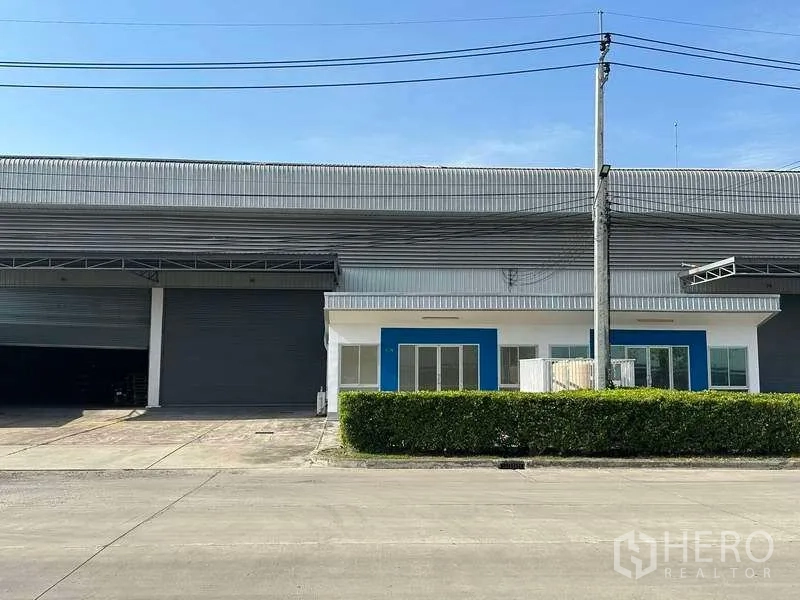工厂 出租在Bang Sao Thong, Samut Prakan - 工厂正面，带大卷帘门与蓝色点缀的办公区，前方有绿篱与车道。