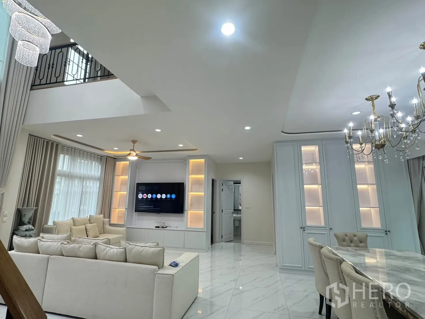 Detached House for rent or sale in Bang Phli, Samut Prakan - Open-plan living and dining area with double-height ceiling, chandelier, built-in TV wall and light sofas on marble-look floors.