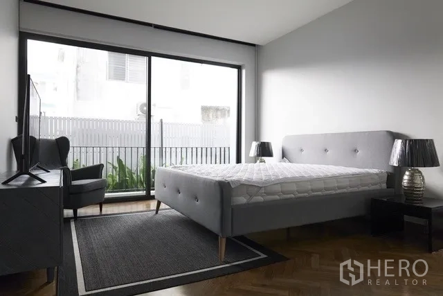 Townhouse for sale in Watthana, Bangkok - Bedroom with gray upholstered bed, armchair and balcony access via sliding door.