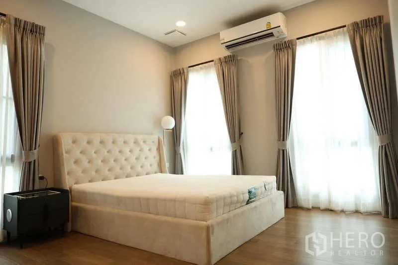 Detached House for rent or sale in Saphan Sung, Bangkok - Corner bedroom with tufted headboard, three tall windows and air conditioner.