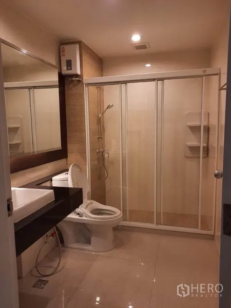 Condominium for rent in Watthana, Bangkok - Bathroom with large mirror, single vanity, toilet and glass-enclosed shower with water heater.