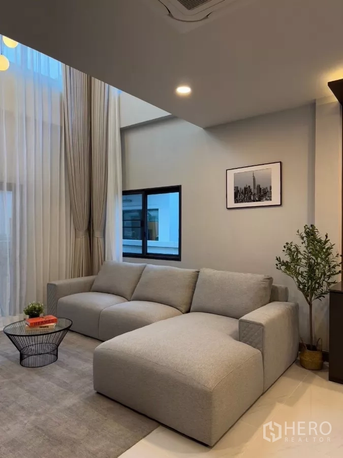 独立式住宅 出租在Bang Phli, Samut Prakan - 客厅双层挑高，落地纱帘与灰色L形沙发