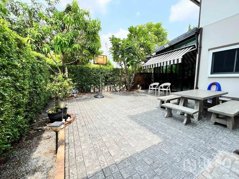 Detached House for sale in Bang Kruai, Nonthaburi - Spacious tiled patio garden with trees, privacy hedge, striped awning, and outdoor seating.