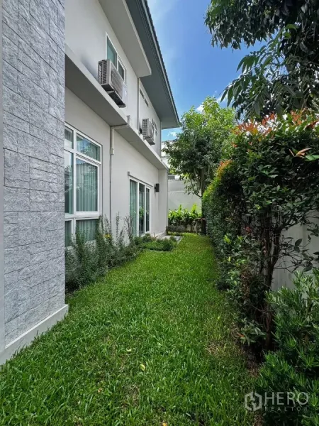 Detached House for rent in Prawet, Bangkok - Side yard lawn beside the two-storey facade with sliding doors and exterior AC units.