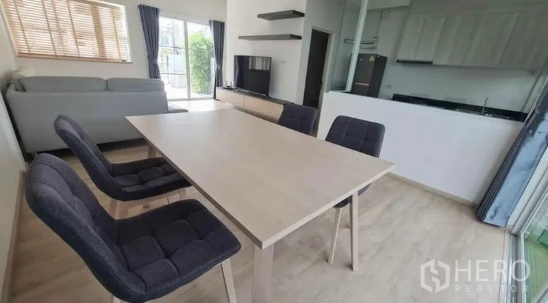 Townhouse for rent in Prawet, Bangkok - Dining space with a four-seat table beside a TV wall and glass partition to the kitchen.