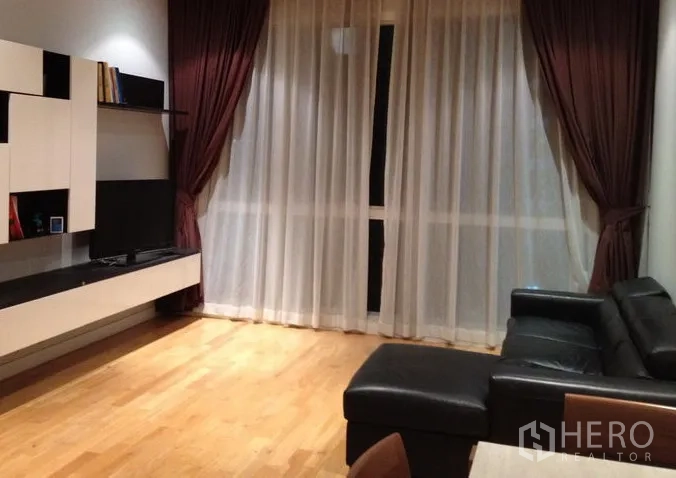 Condominium for rent in Khlong Toei, Bangkok - Modern living room with wood flooring, floor-to-ceiling curtains, wall-mounted media console and black leather sectional sofa in a Khlong Toei condo.
