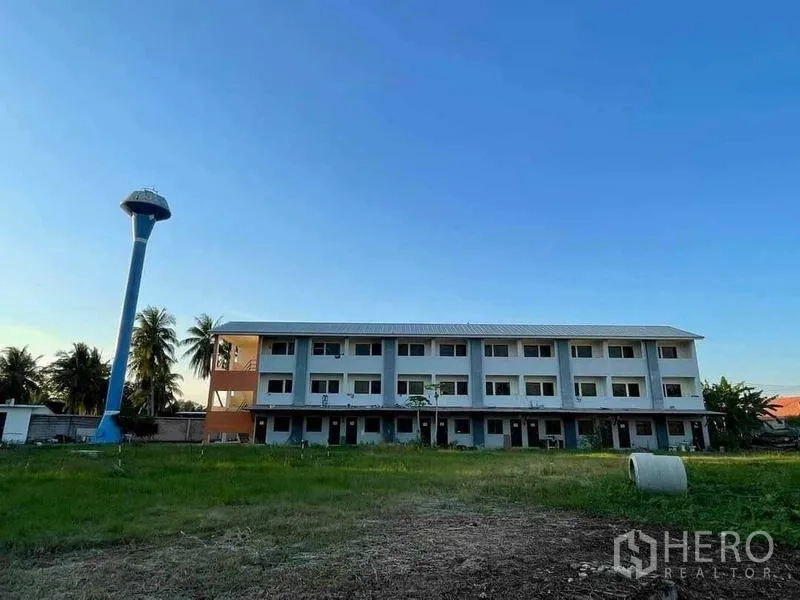 Warehouse for sale in Chon Buri - Three-storey staff dormitory block beside a tall blue water tower and open lawn.