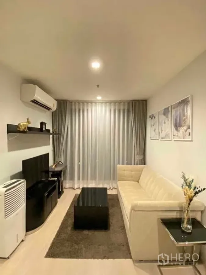 Condominium for rent in Khlong Toei, Bangkok - Bright living room with beige sofa, TV unit, floating shelves and full-height sheer curtains.