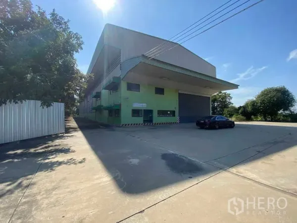 货栈 出租在Lat Lum Kaeo, Pathum Thani - 大型绿色仓库，带挑檐装卸雨棚、卷帘门和宽阔混凝土场地，蓝天之下。