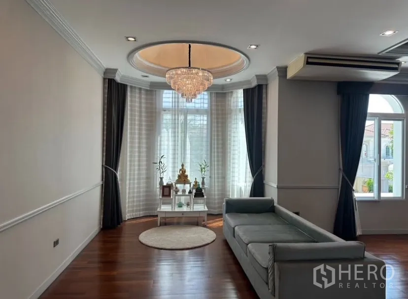 Detached House for rent or sale in Prawet, Bangkok - Lounge area with chandelier, long sofa, and curved bay window dressed with layered curtains.