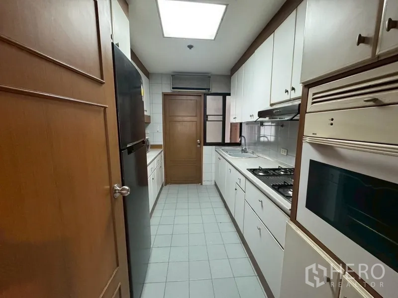 Condominium for rent in Watthana, Bangkok - Long galley kitchen with white cabinets, gas hob, oven, and a large refrigerator.