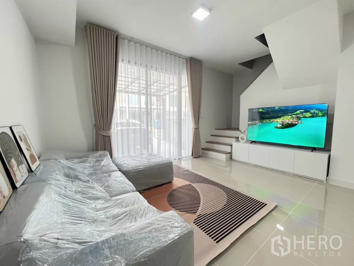 联排别墅 出租在Bang Phli, Samut Prakan - 客厅设落地窗帘，楼梯下方摆放电视
