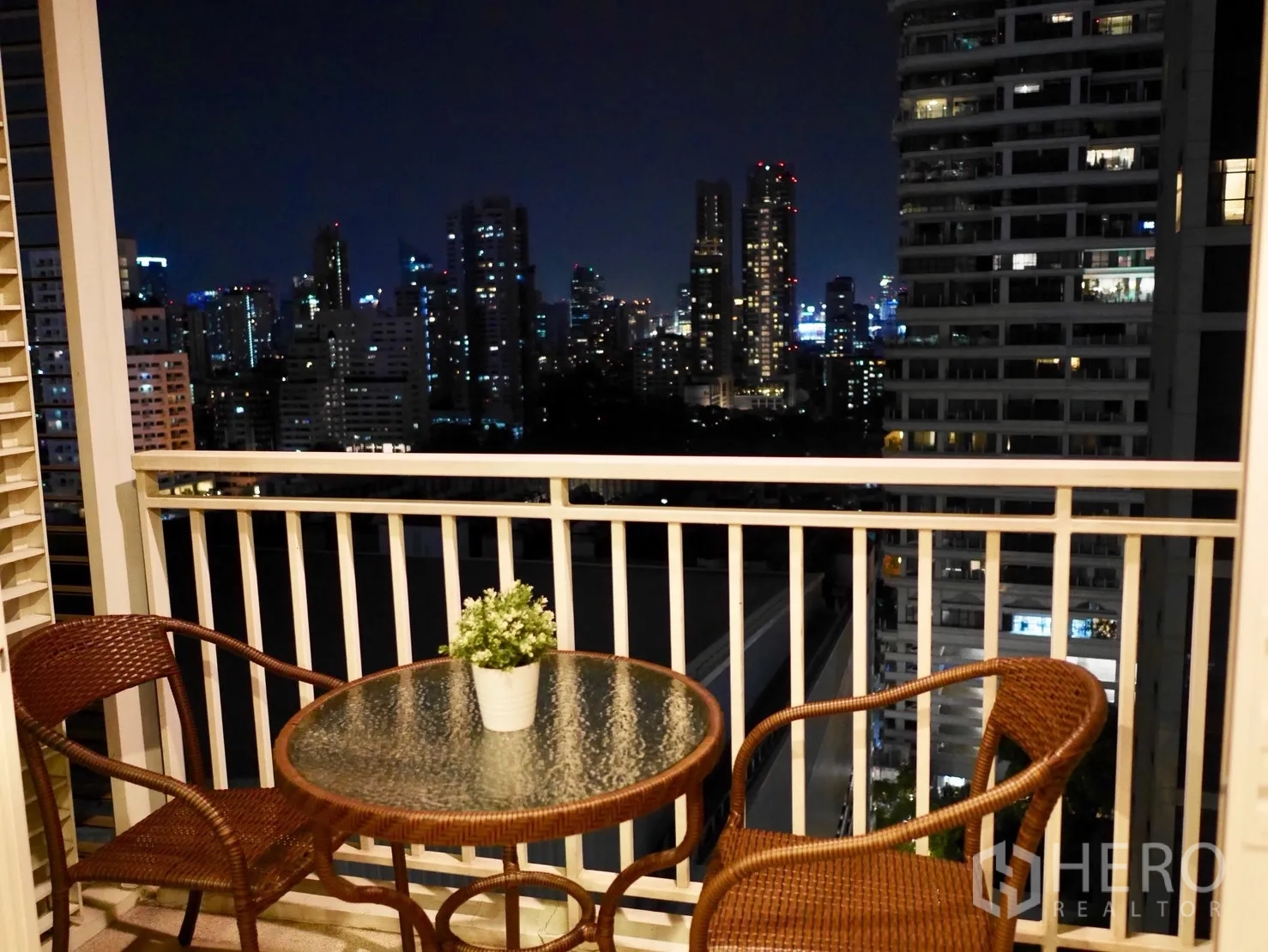 Condominium for rent in Watthana, Bangkok - Balcony with rattan table and chairs overlooking the Bangkok skyline at night.