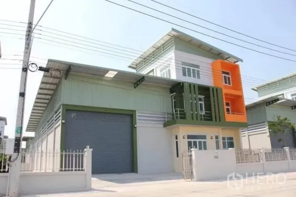 工厂 出售在Lam Luk Ka, Pathum Thani - 兰鲁甲现代化工厂外观，大型卷闸门、彩色办公楼与带围栏车道。