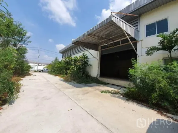 工厂 出租在Lam Luk Ka, Pathum Thani - 工厂外观，宽阔混凝土车道、带顶装卸区与卷闸门，周围绿化，位于Lam Luk Ka。