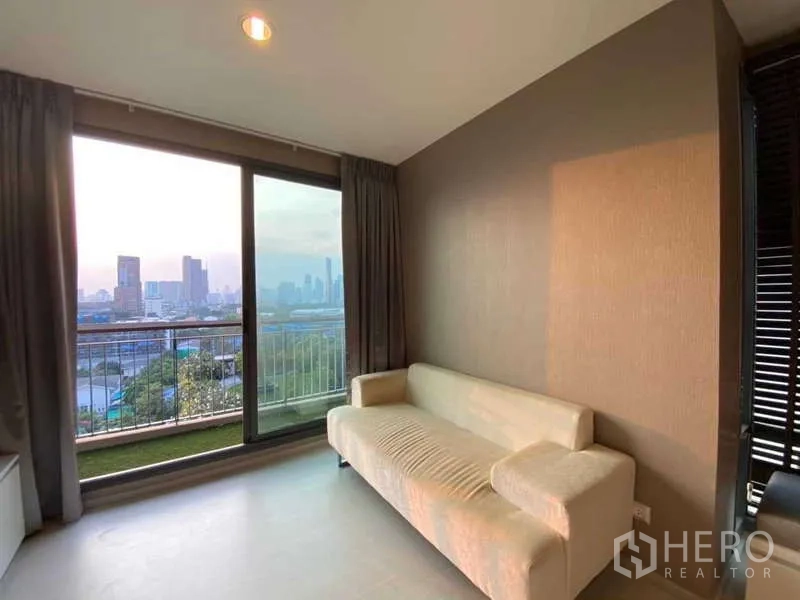 Condominium for rent or sale in Khlong Toei, Bangkok - Living area with large windows and balcony railings overlooking the Bangkok cityscape at dusk.