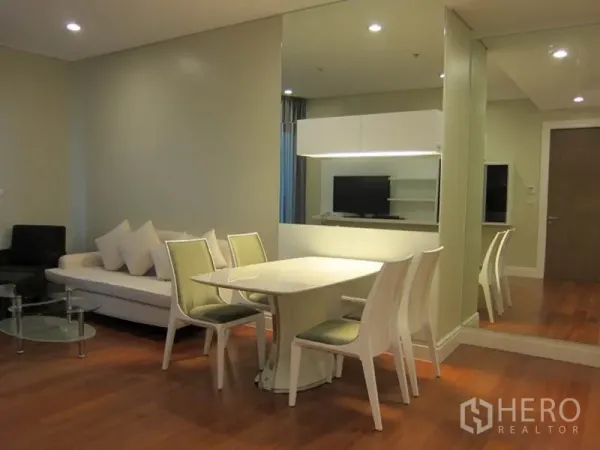 Condominium for rent in Khlong Toei, Bangkok - Open-plan Sukhumvit condo living and dining area with white table, sofa, mirror wall and warm wood flooring.