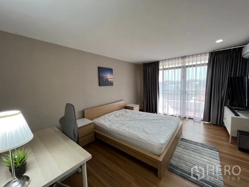 Townhouse for rent or sale in Suan Luang, Bangkok - Cozy bedroom with study desk, balcony access and city outlook through sheer curtains.