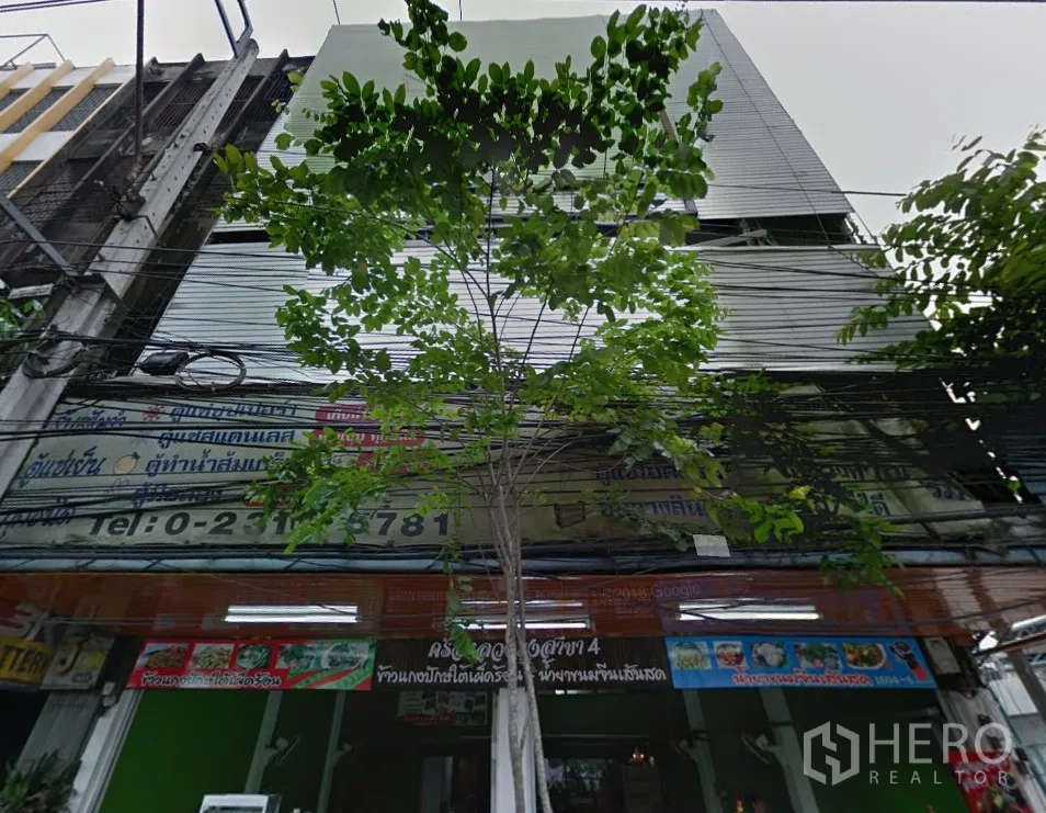 店屋 出租在Phra Khanong, Bangkok - 商铺一排的临街立面，卷闸门与招牌位于树荫下。