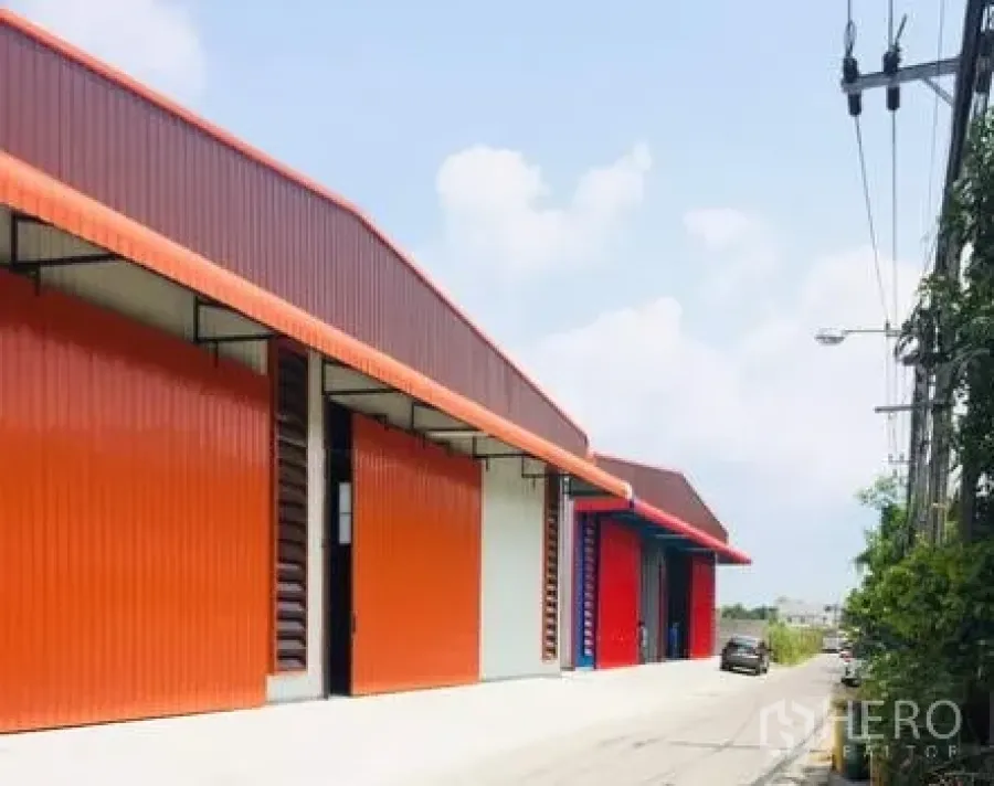 Warehouse for rent in Khan Na Yao, Bangkok - Row of modern orange steel warehouses with large roller doors along a quiet road in Khan Na Yao, Bangkok.