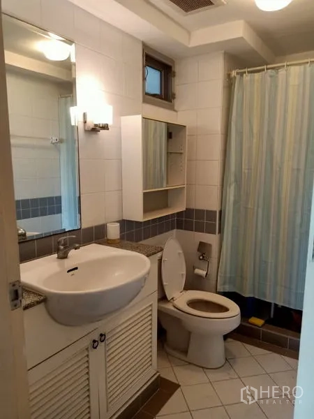 Condominium for rent in Khlong Toei, Bangkok - White-tiled bathroom with vanity sink, mirror cabinet, toilet and a shower with curtain.