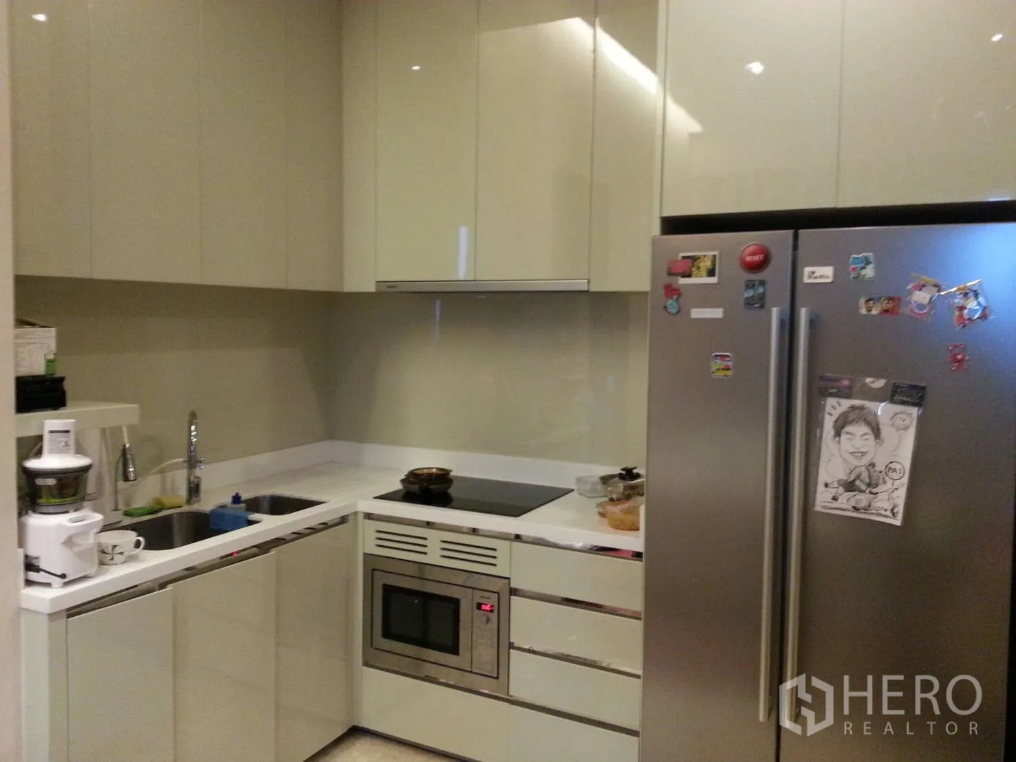 Condominium for rent or sale in Khlong Toei, Bangkok - Modern L-shaped kitchen with glossy cabinets, double sink, induction cooktop, built-in oven and large stainless refrigerator.