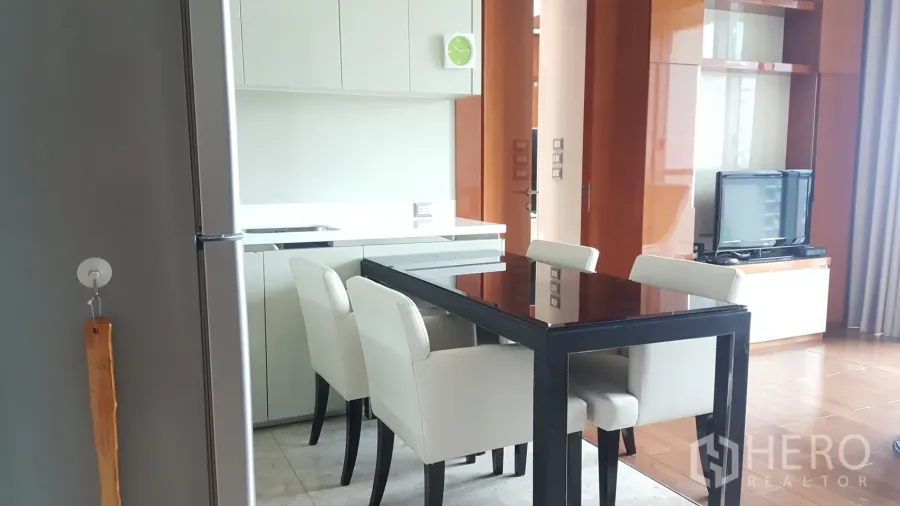 Condominium for rent or sale in Khlong Toei, Bangkok - Open-plan kitchen and dining area with white cabinetry, a black glass-top table and four upholstered chairs, adjoining a living zone with TV unit.