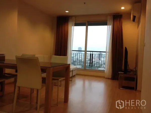Condominium for rent in Huai Khwang, Bangkok - Bright living and dining area with wood flooring, TV console, and balcony offering wide city views through sliding glass doors.