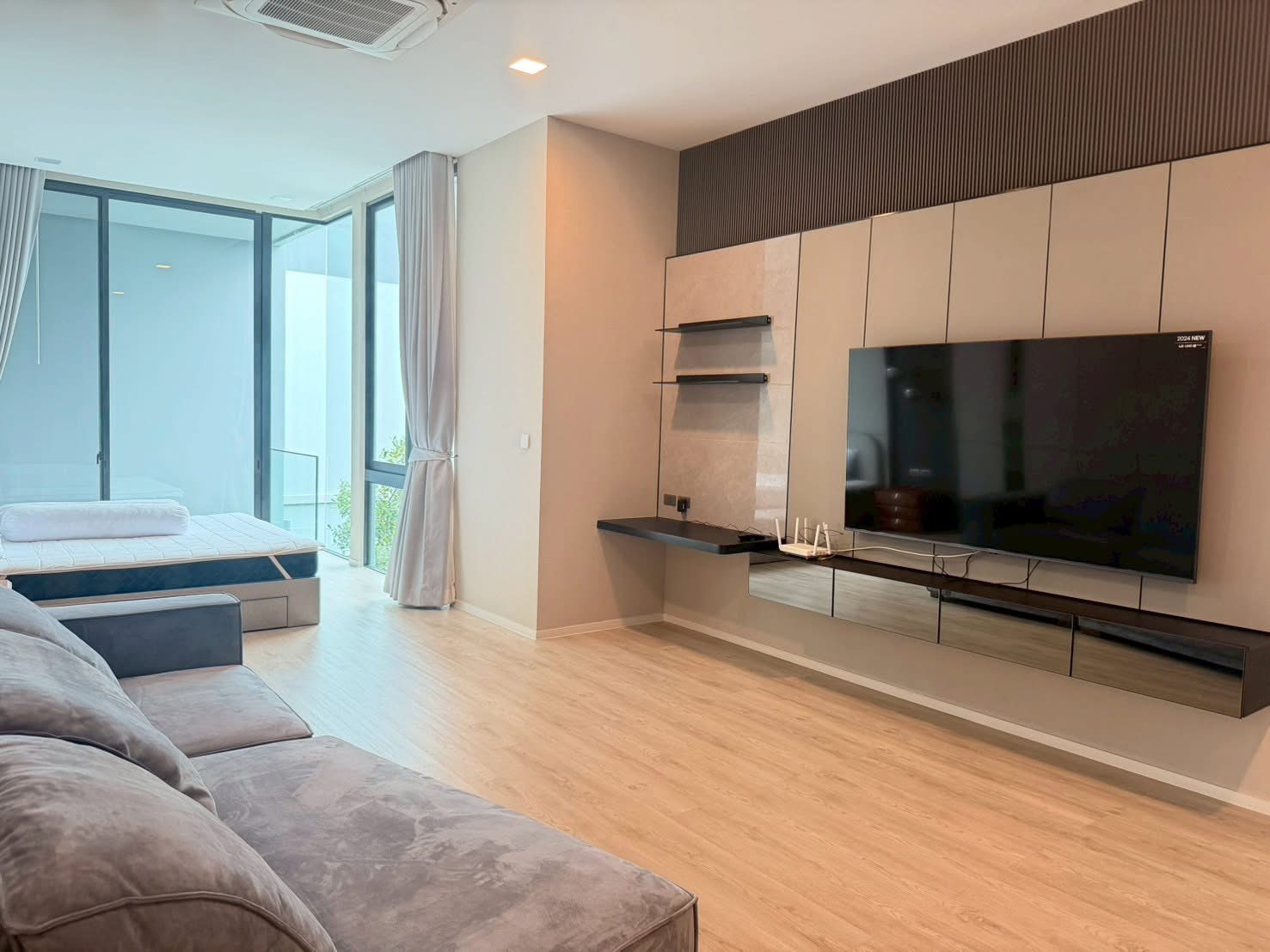 Detached House for rent in Saphan Sung, Bangkok - Spacious family room with built-in media wall, large TV and grey sofa beside a bedroom area and tall windows.