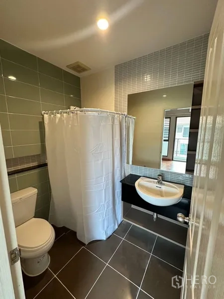 Townhouse for rent or sale in Phra Khanong, Bangkok - Compact bathroom with shower curtain, large mirror, and green wall tiles.