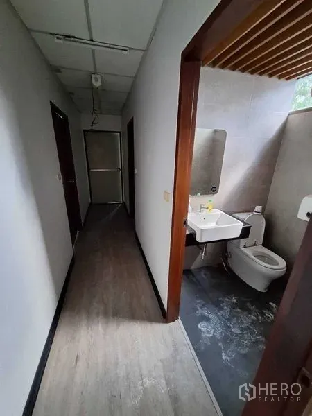 办公空间 出租在Watthana, Bangkok - 卫生间带洗手台、镜子、坐便器与灰色瓷砖，紧邻走廊。