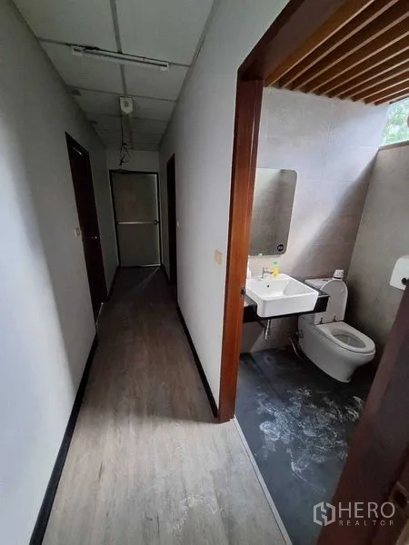 办公空间 出租在Watthana, Bangkok - 卫生间带洗手台、镜子、坐便器与灰色瓷砖，紧邻走廊。