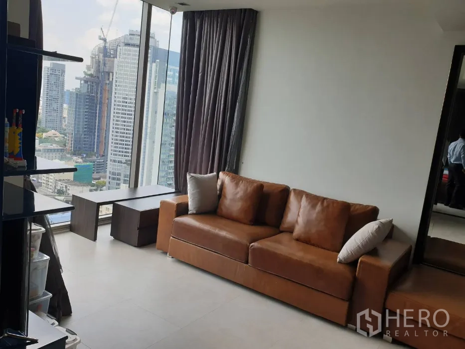Condominium for rent in Khlong Toei, Bangkok - Bright living area with floor-to-ceiling windows, city skyline views, and a brown sofa beside dark curtains.