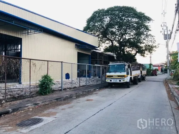 Warehouse for rent in Wang Thonglang, Bangkok - Warehouse exterior along a quiet access road with a parked flatbed truck and a large shade tree.