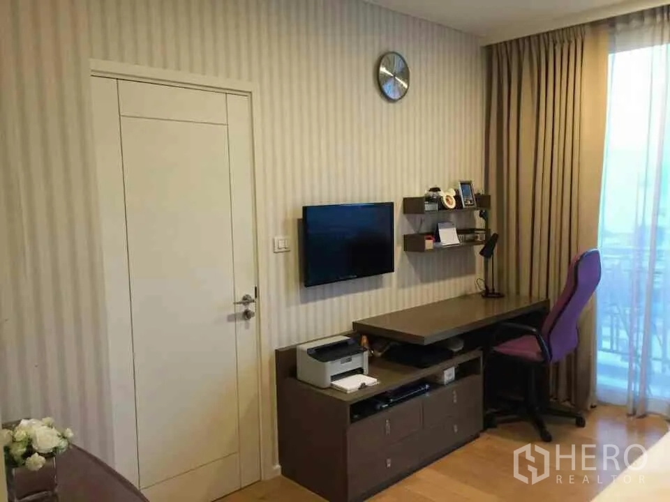 Condominium for rent or sale in Watthana, Bangkok - Work-from-home corner with desk, wall-mounted TV, shelving and curtains leading to a balcony.