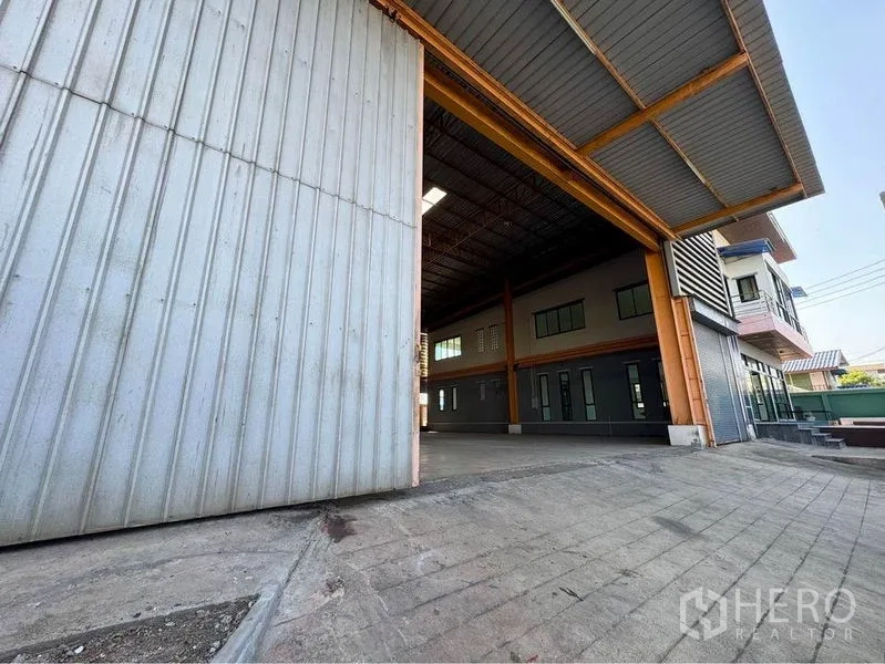 Warehouse for rent or sale in Mueang Samut Sakhon, Samut Sakhon - Exterior warehouse entry showing a large steel door, ramped driveway, and adjoining office facade.