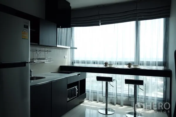 Condominium for rent or sale in Khlong Toei, Bangkok - Bright kitchen with dark cabinetry, built‑in appliances and a breakfast bar with two stools by floor‑to‑ceiling windows.