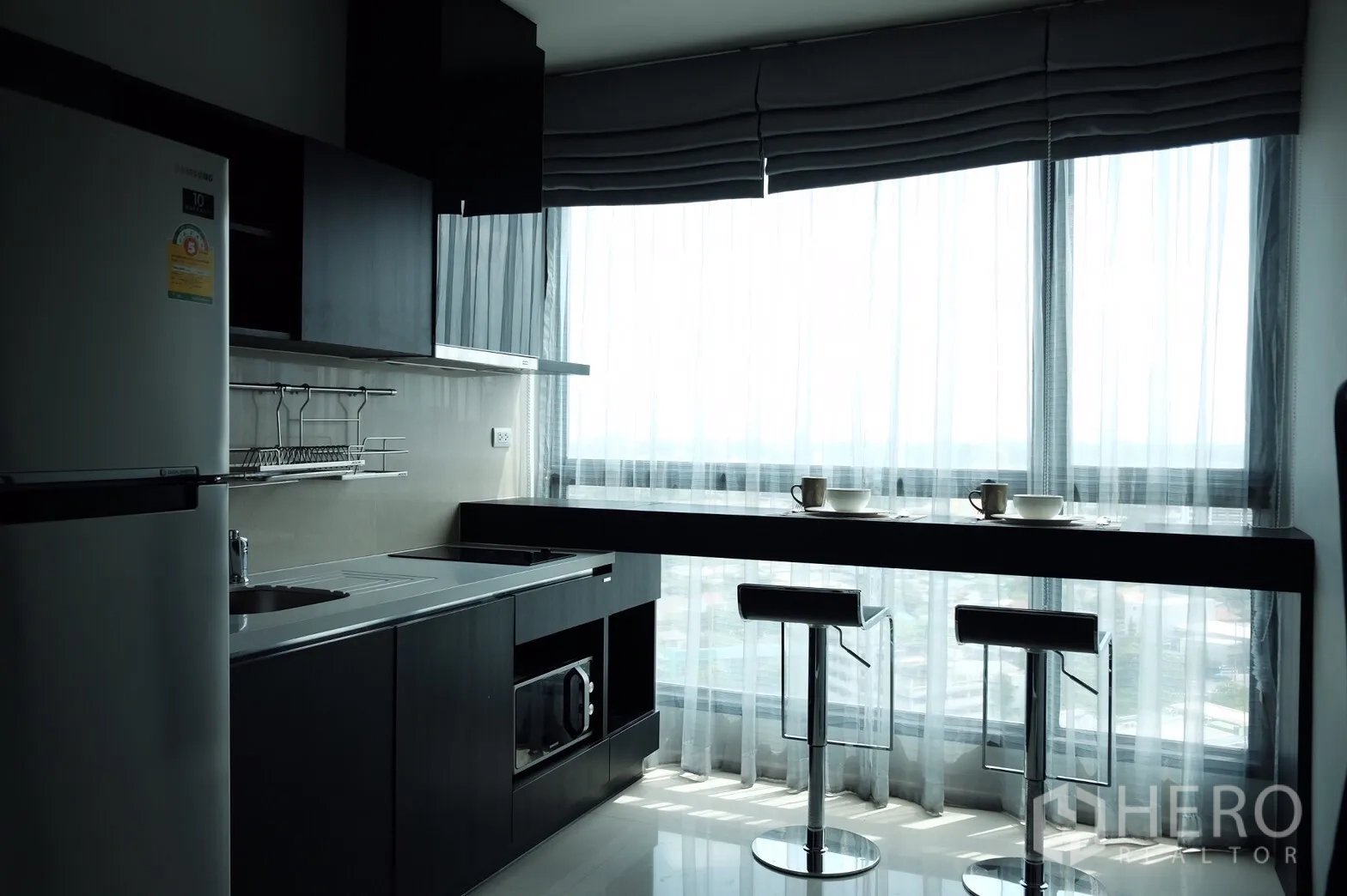 Condominium for rent or sale in Khlong Toei, Bangkok - Bright kitchen with dark cabinetry, built‑in appliances and a breakfast bar with two stools by floor‑to‑ceiling windows.
