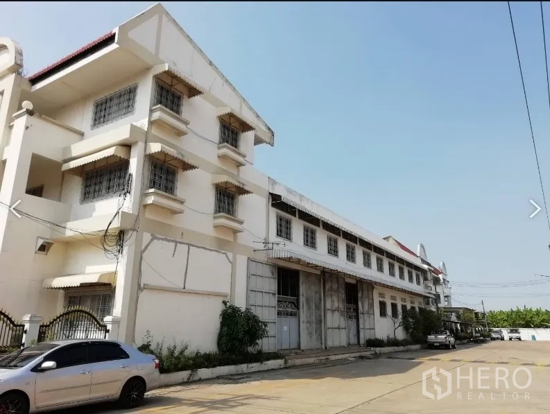 Warehouse for rent in Nakhon Pathom - Street view of a large multi-storey white warehouse with gated frontage, wide driveway and loading doors in an industrial lane.