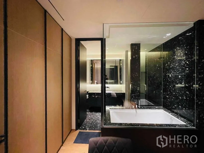 Condominium for rent in Khlong Toei, Bangkok - Black marble bathroom with built-in bathtub behind clear glass and ambient lighting.