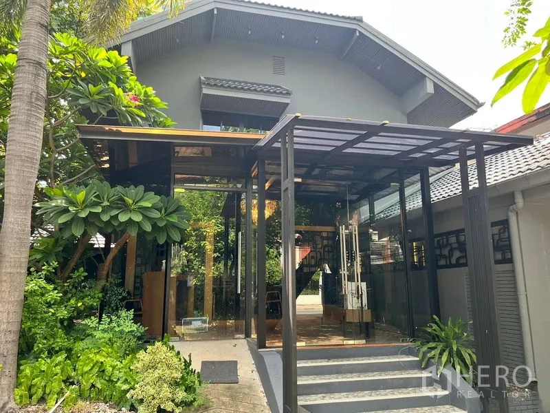 Retail Space for rent in Bangkok - Modern glass entrance pavilion framed by greenery beside a pitched-roof building.