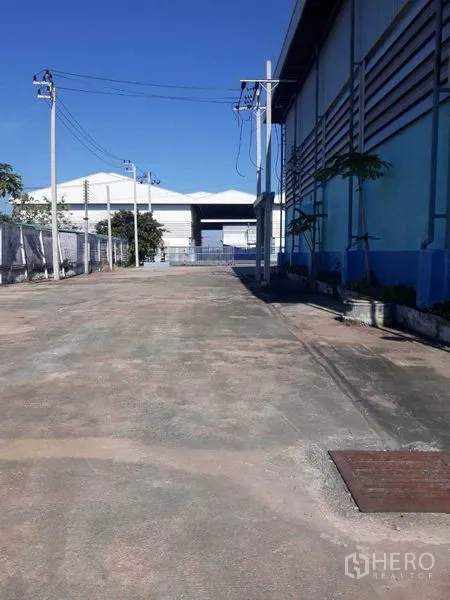Factory for rent in Samut Prakan - Wide driveway beside the factory with power poles and fenced boundary under clear sky.