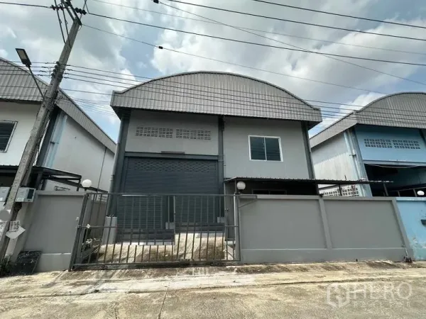 货栈 出租在Pak Kret, Nonthaburi - 仓库正立面带围栏与宽幅卷闸门，弧形金属屋顶，位于帕克烈。