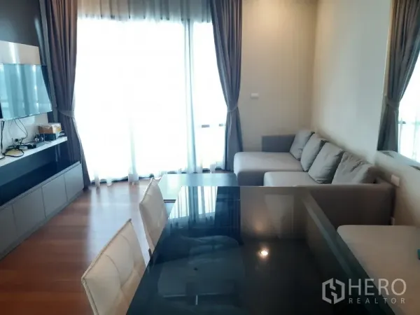 Condominium for rent or sale in Khlong Toei, Bangkok - Bright living and dining area with L-shaped sofa, TV wall, floor-to-ceiling curtains and a glass table.