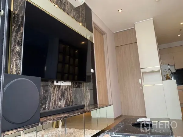 Condominium for sale - Living room media wall with large TV and marble finish facing compact kitchen in Bangkok condo.