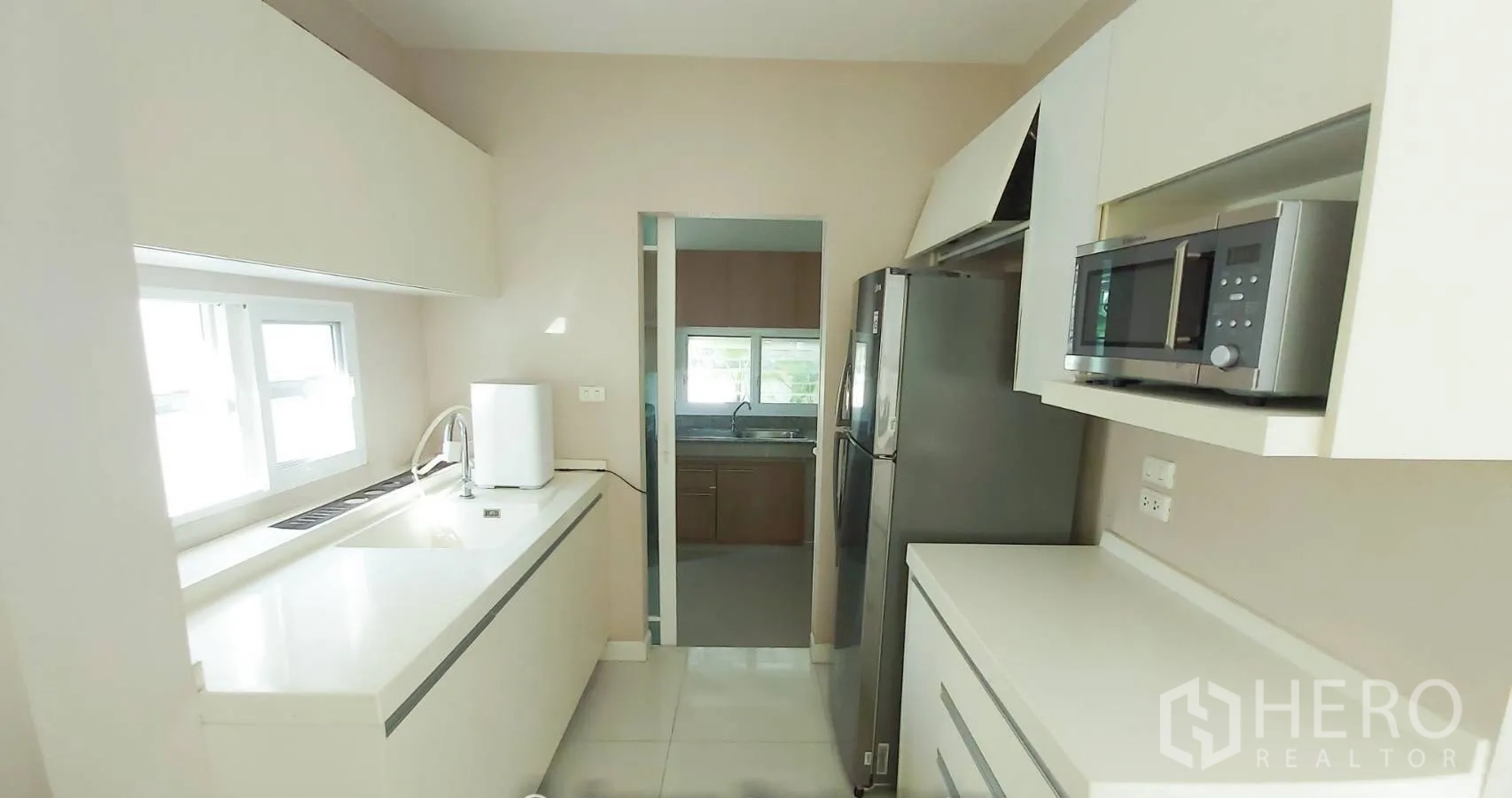 Detached House for sale in Bang Kapi, Bangkok - Bright galley kitchen with white countertops, built-in cabinets and window, opening to a Thai kitchen.