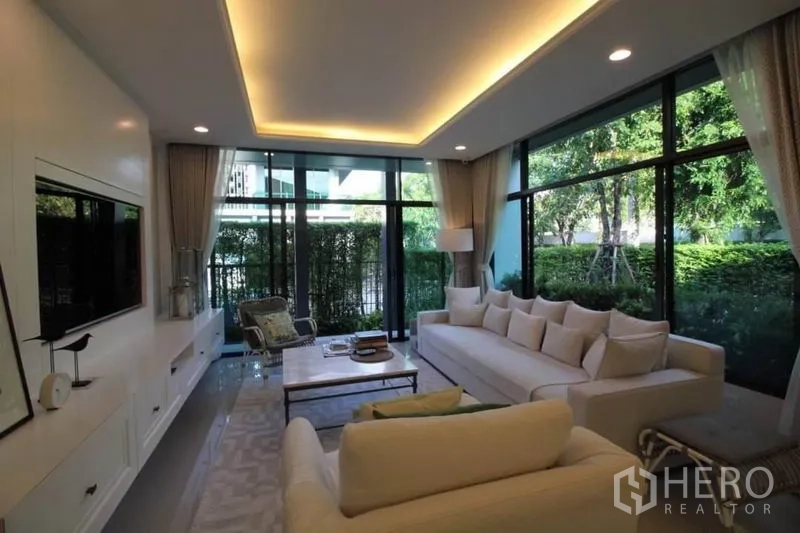 Detached House for rent or sale in Prawet, Bangkok - Sunlit living room with white sofas, TV wall, cove lighting, and expansive glass overlooking hedges.